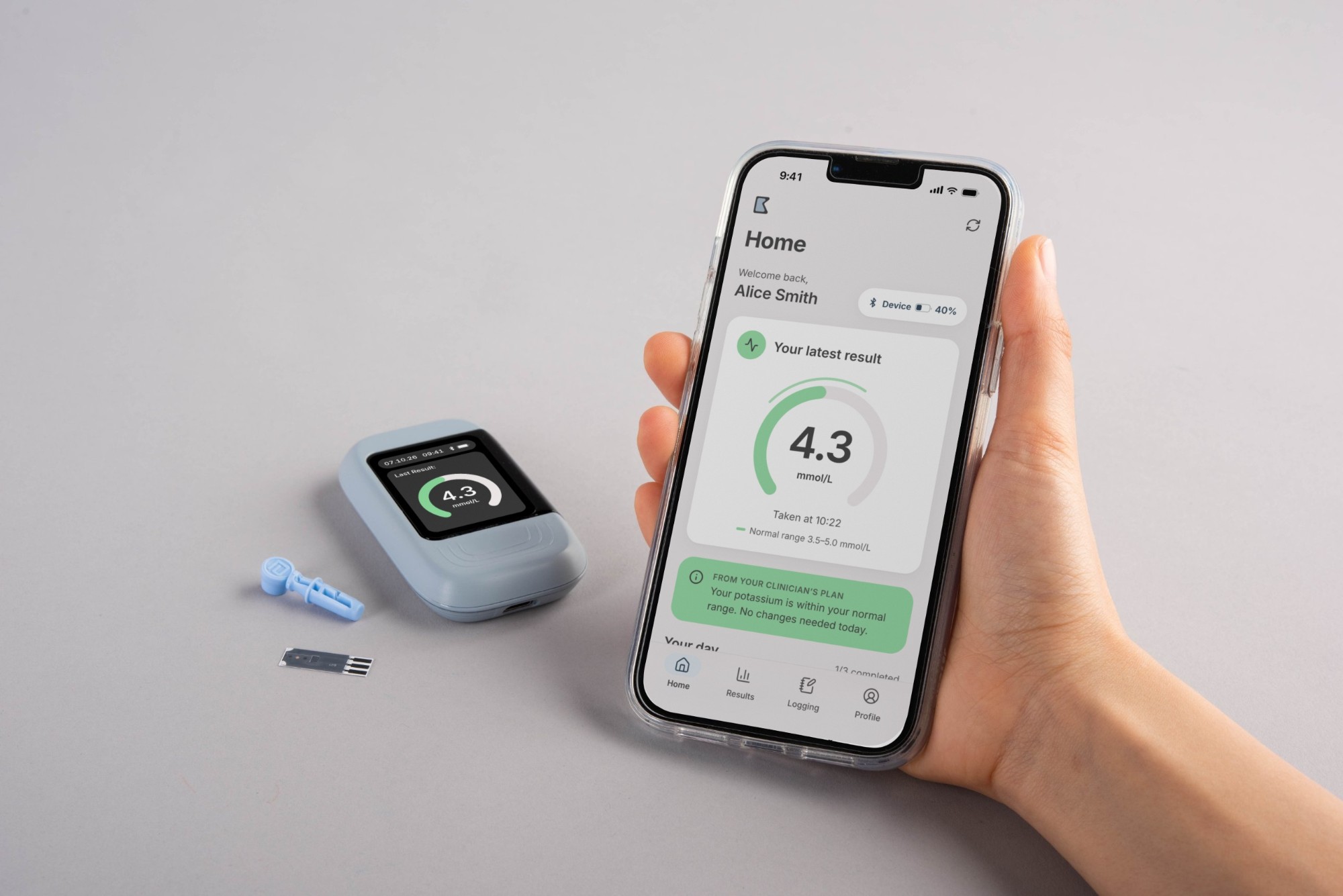The complete Kali system: the monitor, a lancet and test strip on a table, and a hand holding a phone showing the app's latest result.